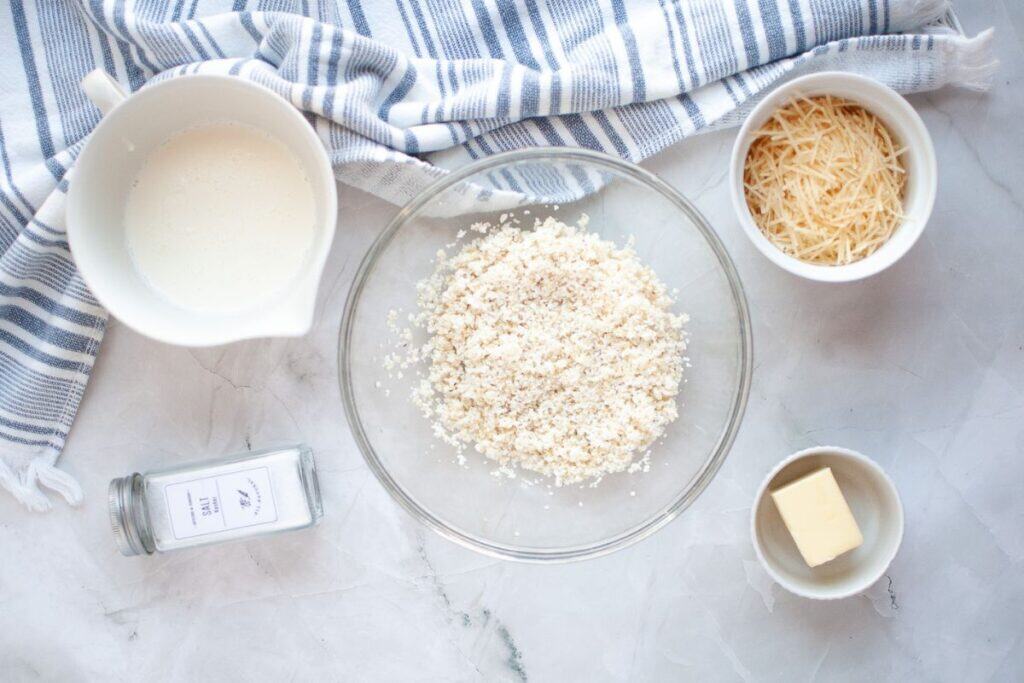A bowl of breadcrumbs, grated cheese, butter, cream, and a jar of salt on a marble surface with a striped cloth.