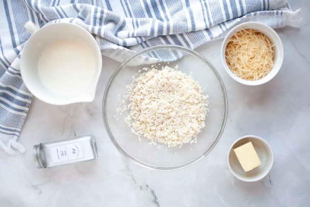 A bowl of breadcrumbs, grated cheese, butter, cream, and a jar of salt on a marble surface with a striped cloth.