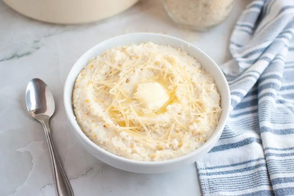 A bowl of grits topped with shredded cheese and a pat of butter, next to a spoon and a striped dish towel on a marble surface.