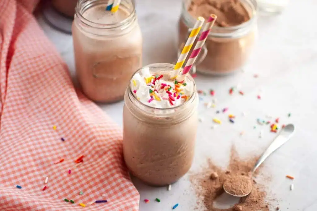 A close-up, top-down shot of a clear glass filled with white whipped cream, generously topped with colorful star-shaped sprinkles, covering a light brown frozen hot chocolate.