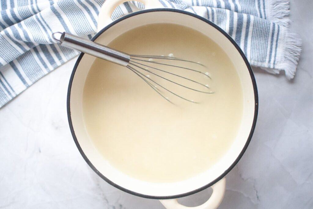 A whisk rests in a cream-colored sauce inside a white pot with black trim, set on a white marble surface next to a blue and white striped towel.