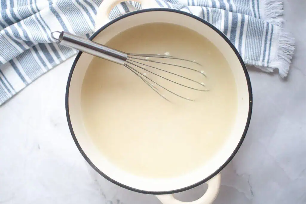 A whisk rests in a cream-colored sauce inside a white pot with black trim, set on a white marble surface next to a blue and white striped towel.