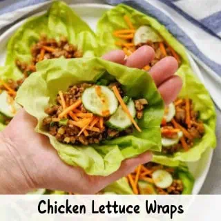 A hand holding a chicken lettuce wrap filled with ground chicken, shredded carrots, cucumber slices, and herbs; several more wraps are on a plate in the background.
