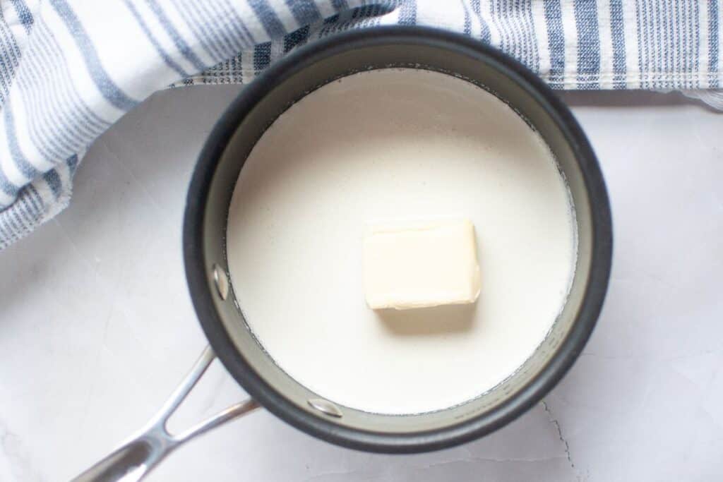 A saucepan containing milk and a block of butter, next to a striped kitchen towel on a white surface.