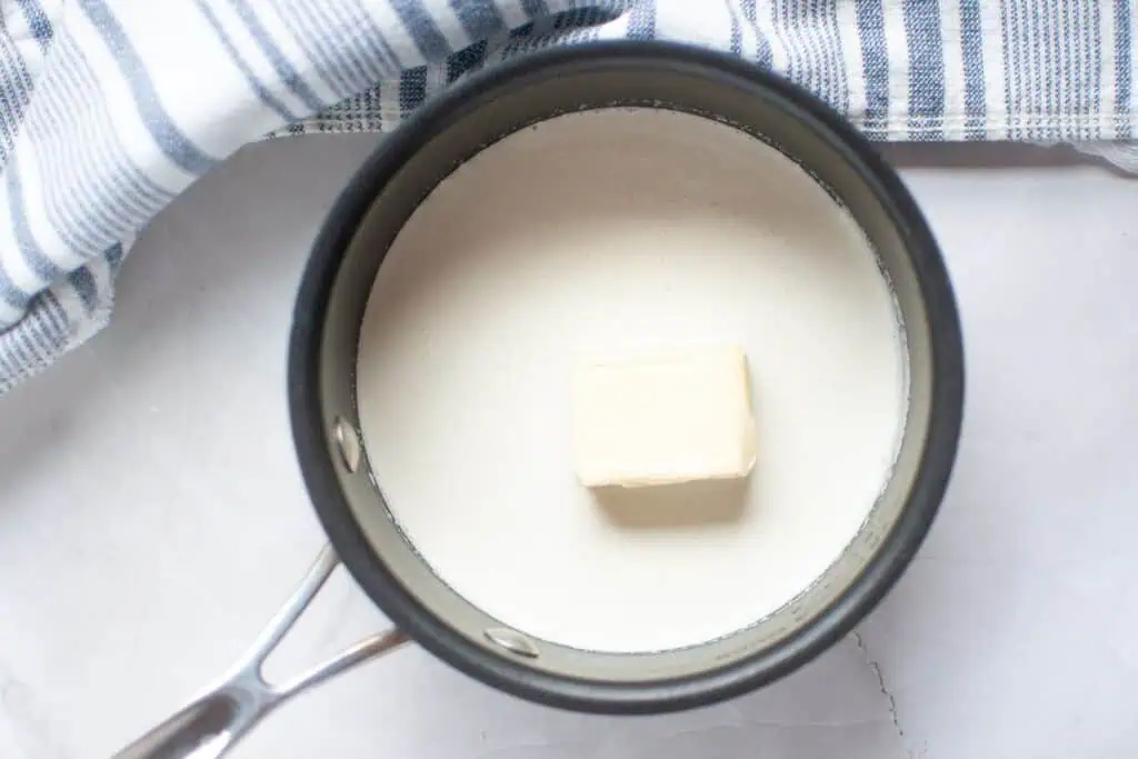 A saucepan containing milk and a block of butter, next to a striped kitchen towel on a white surface.