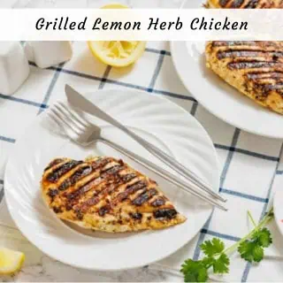 A grilled chicken breast with grill marks is served on a white plate with a knife and fork, garnished with lemon and cilantro on a checkered tablecloth.