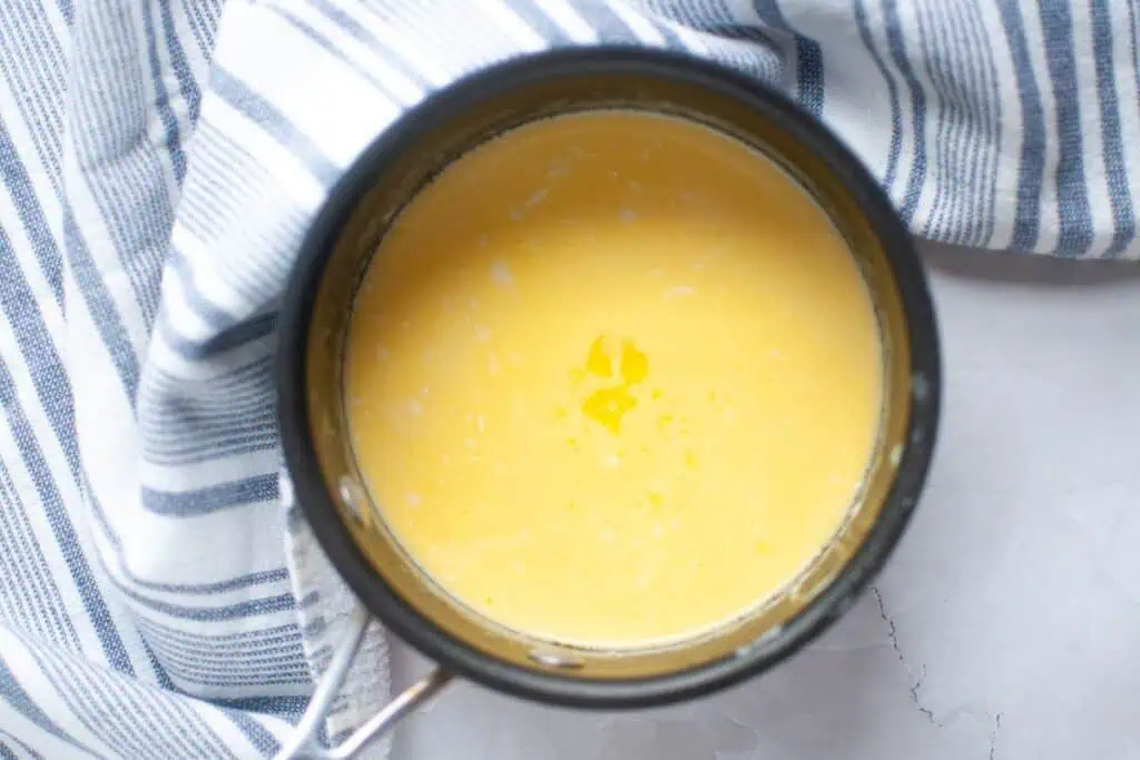 A pot of smooth yellow sauce or custard sits on a white surface next to a blue and white striped towel.