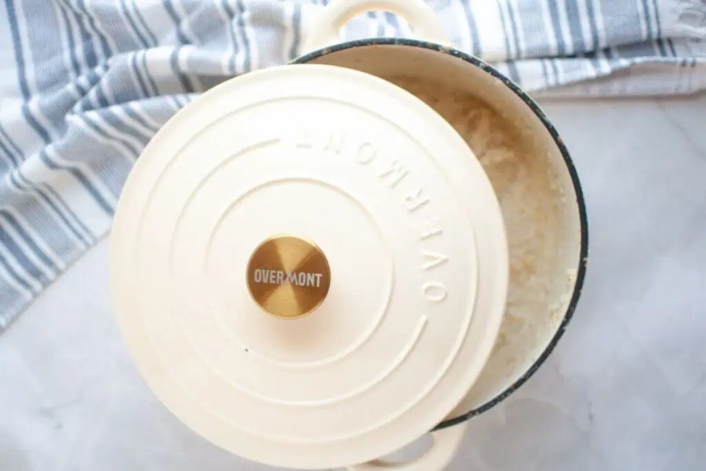 A cream-colored Overmont Dutch oven with a partially open lid reveals cooked rice inside, placed on a white surface with a blue and white striped cloth nearby.