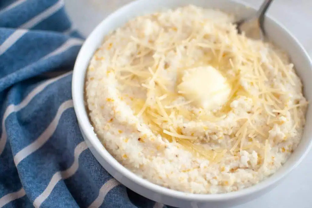 A bowl of creamy grits topped with shredded cheese and a pat of butter, with a spoon and a blue striped cloth nearby.