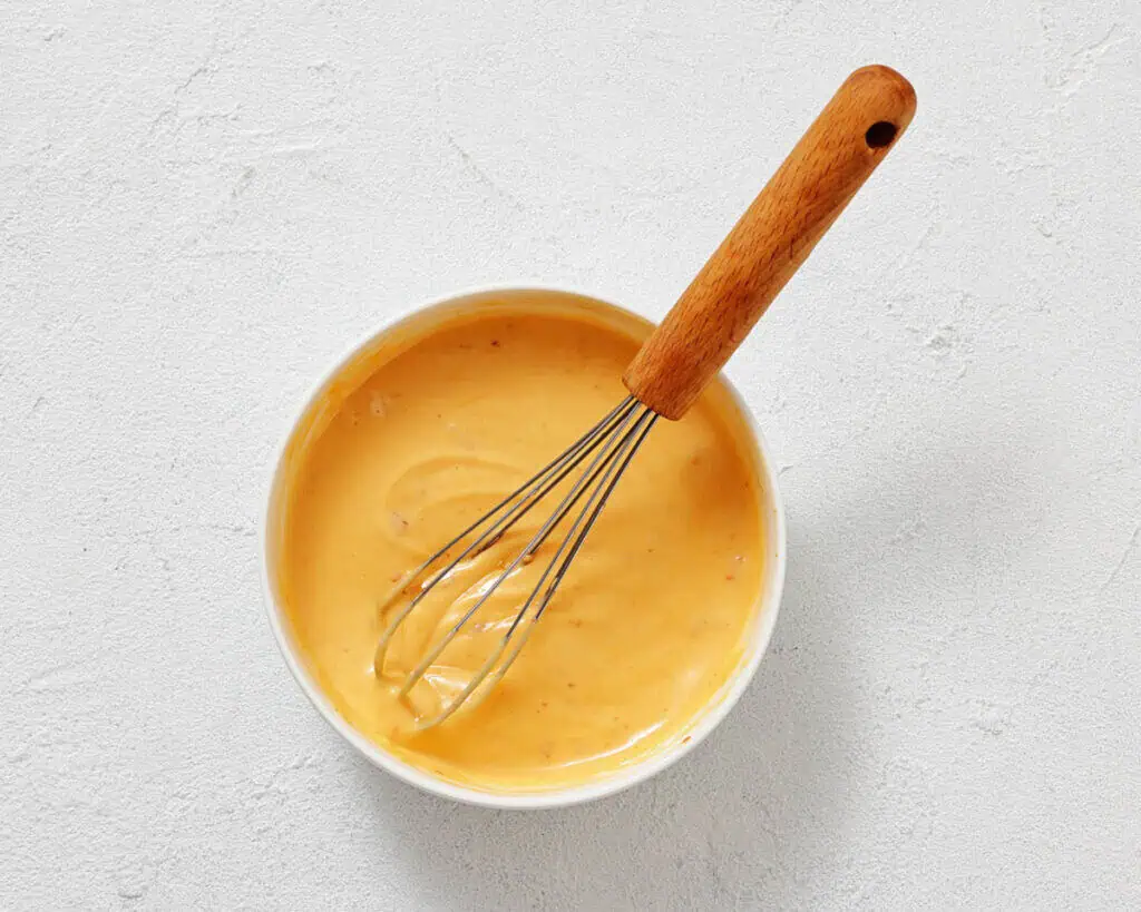 A bowl of creamy orange sauce being whisked with a metal whisk that has a wooden handle, set on a white textured surface.