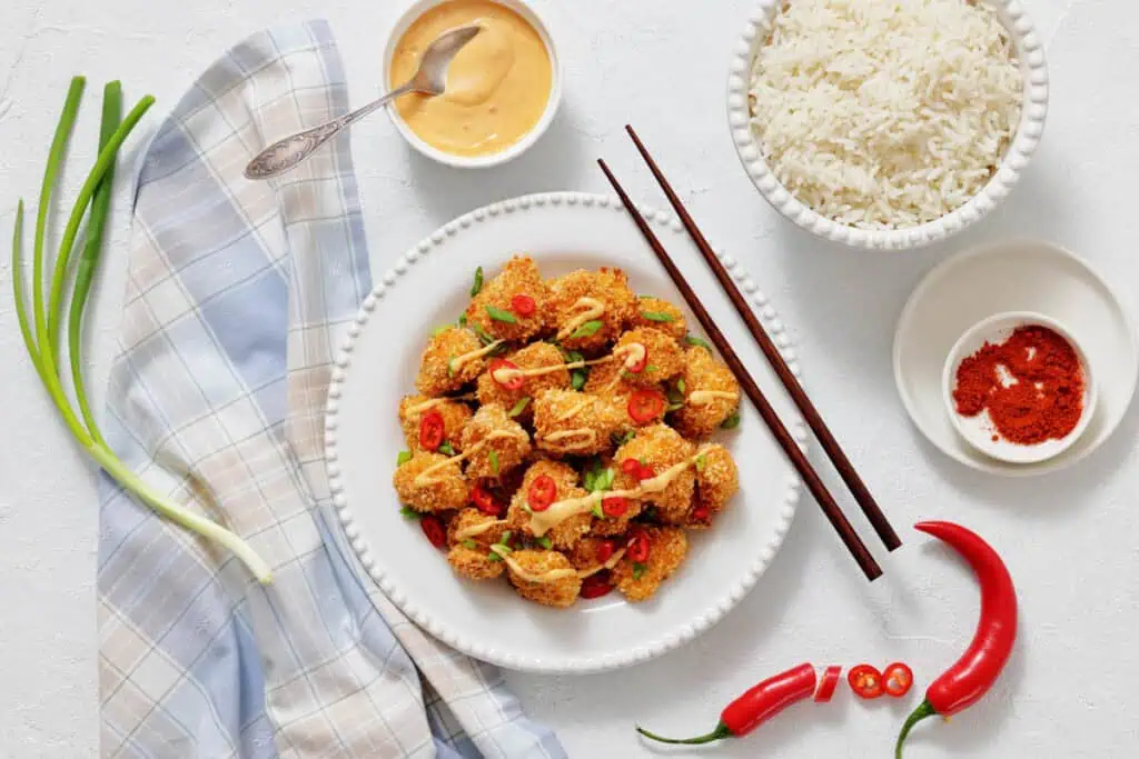 A plate of breaded tofu pieces topped with sliced red chili and green onions, served with white rice, sauce, and spices. Chopsticks and fresh chili peppers are on the side.