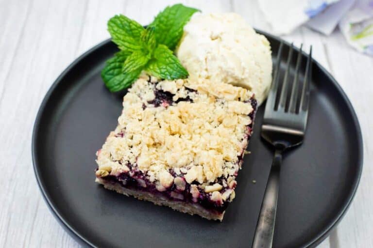 A dessert plate with a berry crumble bar, a scoop of vanilla ice cream, a sprig of mint, and a black fork on a black plate.