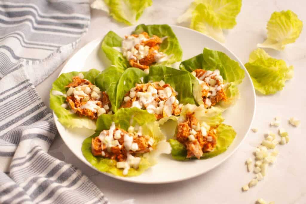 A white plate with six lettuce cups filled with shredded chicken and topped with diced onions and creamy sauce, on a marble surface with a striped towel and lettuce leaves nearby.