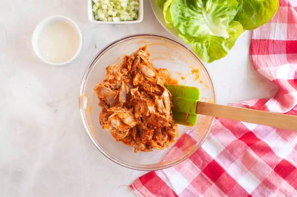A glass bowl of shredded chicken mixed with sauce, a spatula, lettuce leaves, diced cucumbers, dressing, and a red-and-white checkered cloth on a white surface.