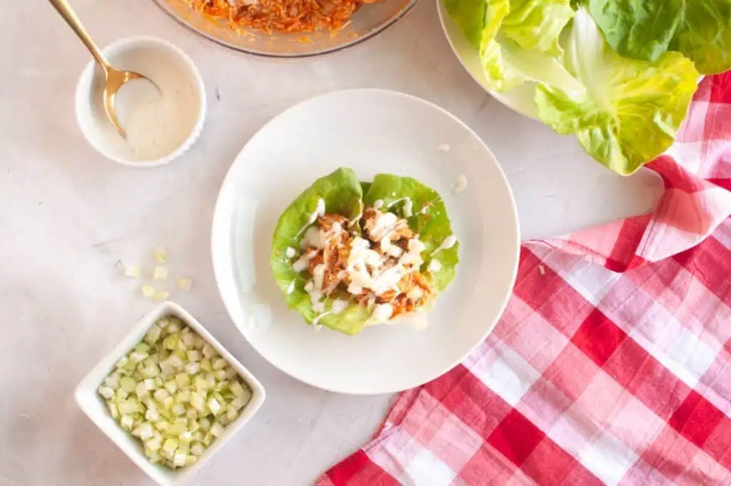 A plate with two lettuce wraps filled with shredded chicken and drizzled with white sauce, surrounded by a bowl of lettuce, diced celery, and a red checkered napkin.