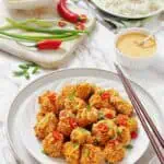 A plate of crispy, breaded tofu bites garnished with sliced red chili and green onions, served with a bowl of white rice, sauce, and fresh vegetables on the side.