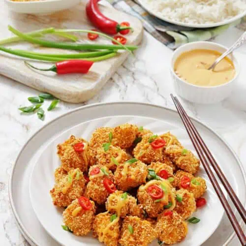 A plate of crispy, breaded tofu bites garnished with sliced red chili and green onions, served with a bowl of white rice, sauce, and fresh vegetables on the side.