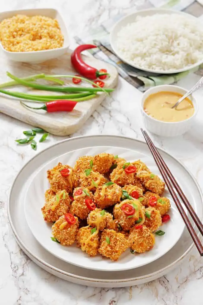 A plate of crispy, breaded tofu bites garnished with sliced red chili and green onions, served with a bowl of white rice, sauce, and fresh vegetables on the side.