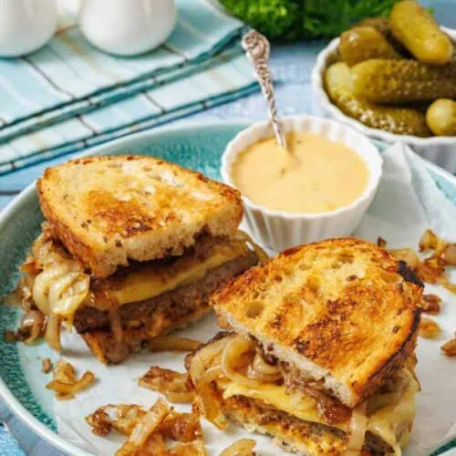A plate with two halves of a grilled sandwich filled with melted cheese and caramelized onions, served with a side of pickles and dipping sauce.