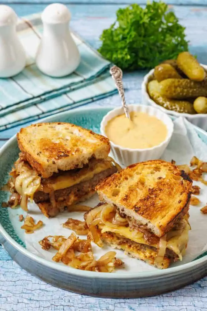 A plate with two halves of a grilled sandwich filled with melted cheese and caramelized onions, served with a side of pickles and dipping sauce.