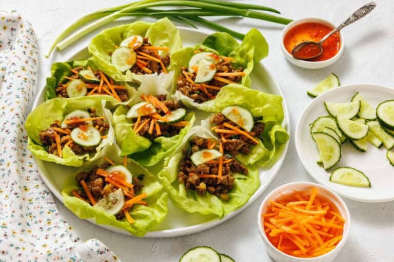 A plate of lettuce wraps filled with cooked ground meat, shredded carrots, and cucumber slices, surrounded by bowls of sliced cucumbers, shredded carrots, dipping sauce, and green onions.