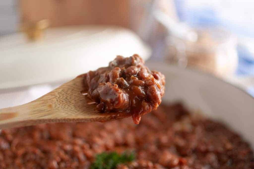A close-up of a wooden spoon holding a serving of baked beans casserole, with a pot of chili in the background.