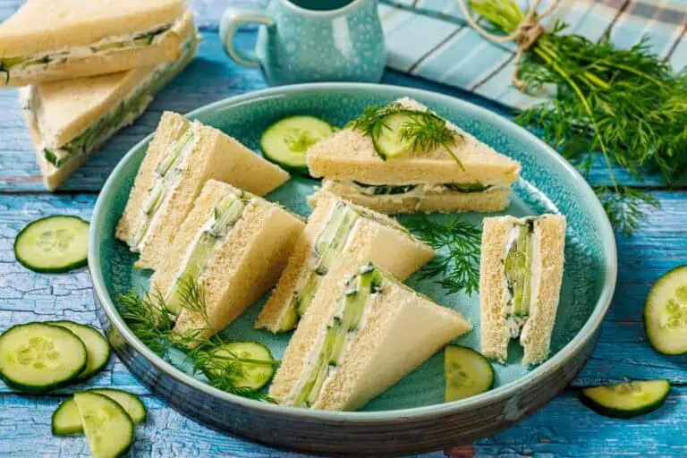 A plate with neatly arranged cucumber sandwiches on white bread, garnished with fresh dill and cucumber slices, sits on a blue wooden table.
