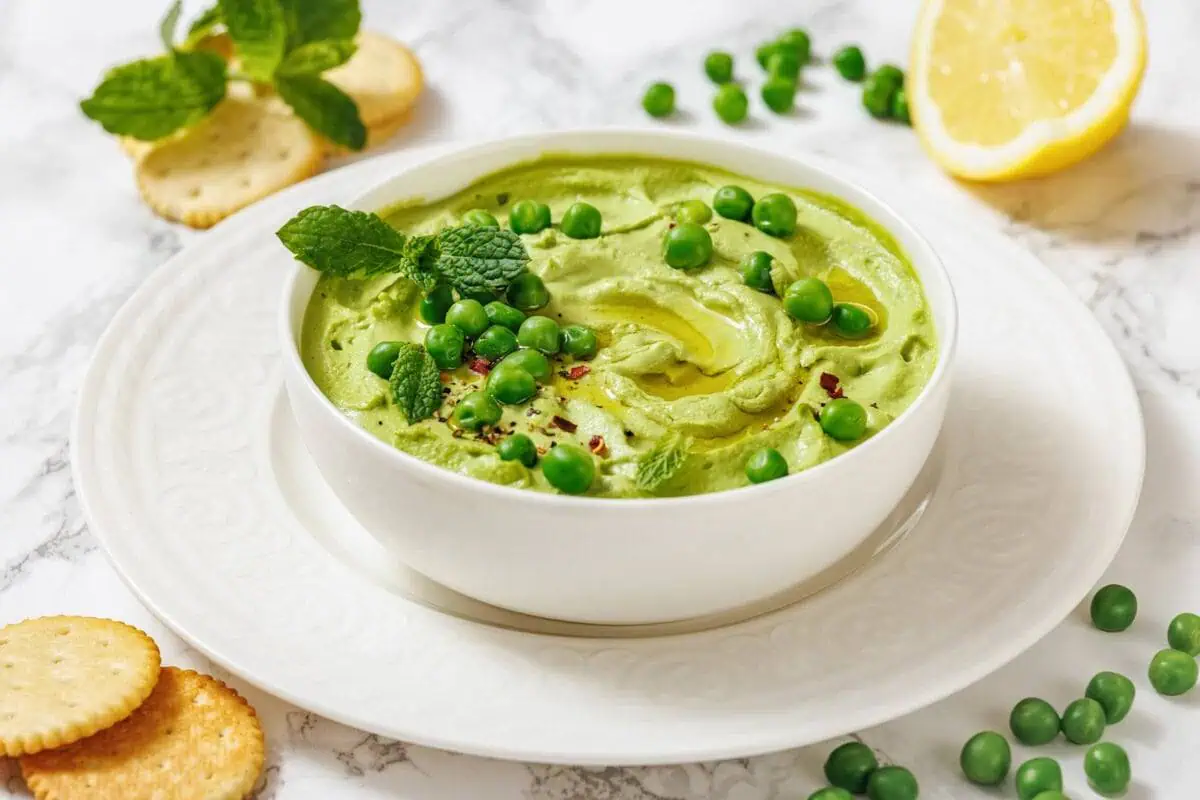 A white bowl of vibrant green Green Pea Hummus, garnished with peas and mint, served with crackers and a lemon wedge.