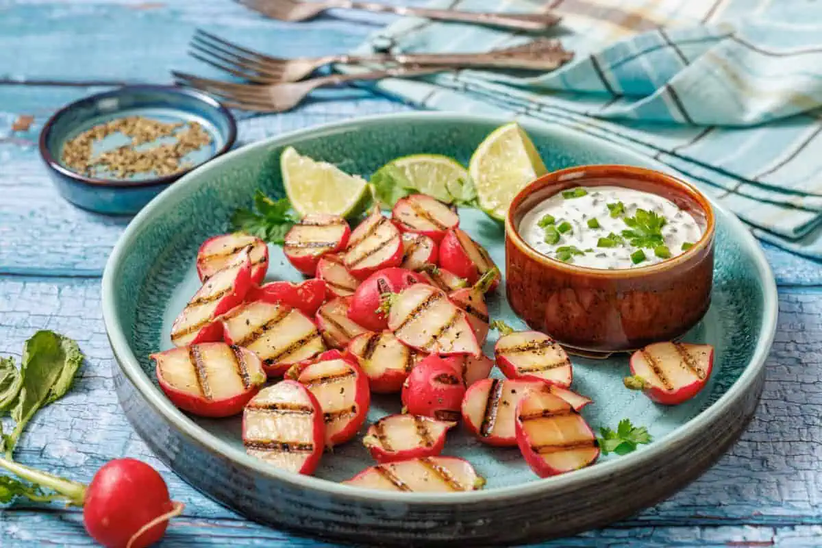 Grilled radish halves on a green plate with lime wedges and a bowl of herb dip; herbs and utensils in the background.