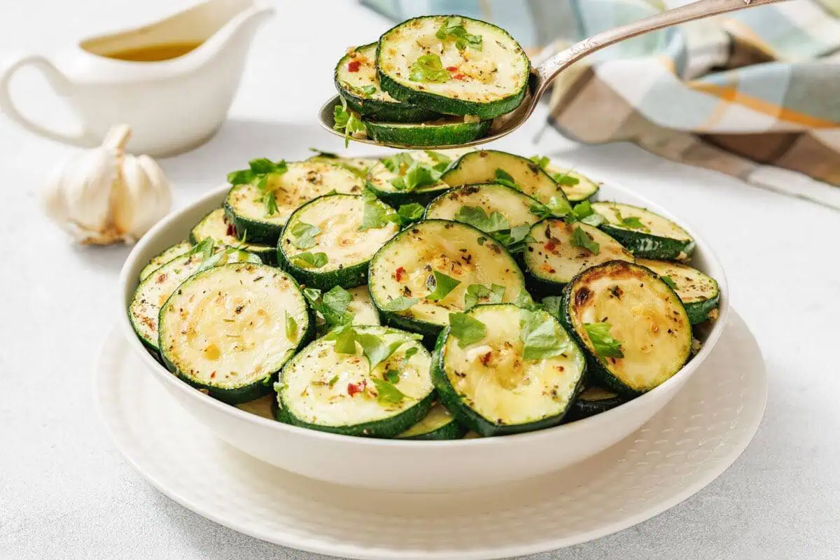 A bowl of sliced, roasted zucchini garnished with herbs, with a spoon serving a portion. A garlic bulb and a jug of sauce are in the background.