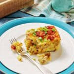 A slice of breakfast casserole topped with bacon and green onions on a white plate with a fork, set on blue and white dishes with a napkin and milk jug in the background.