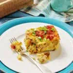 A slice of breakfast casserole topped with bacon and green onions on a white plate with a fork, set on blue and white dishes with a napkin and milk jug in the background.