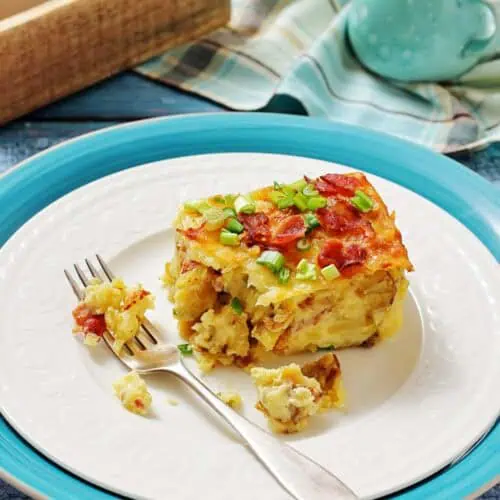 A slice of breakfast casserole topped with bacon and green onions on a white plate with a fork, set on blue and white dishes with a napkin and milk jug in the background.