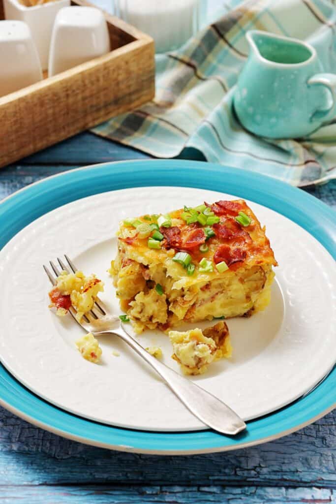 A slice of breakfast casserole topped with bacon and green onions on a white plate with a fork, set on blue and white dishes with a napkin and milk jug in the background.