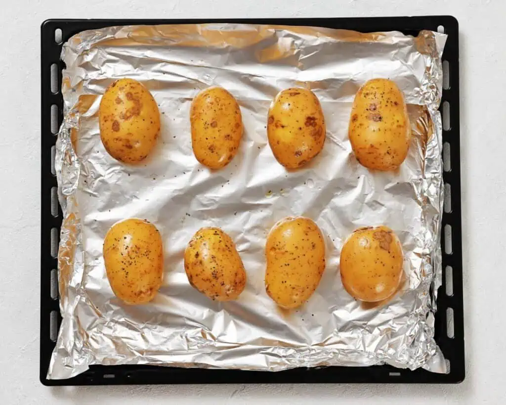 Eight whole, unpeeled potatoes seasoned with pepper are arranged on a foil-lined baking tray, ready for roasting.