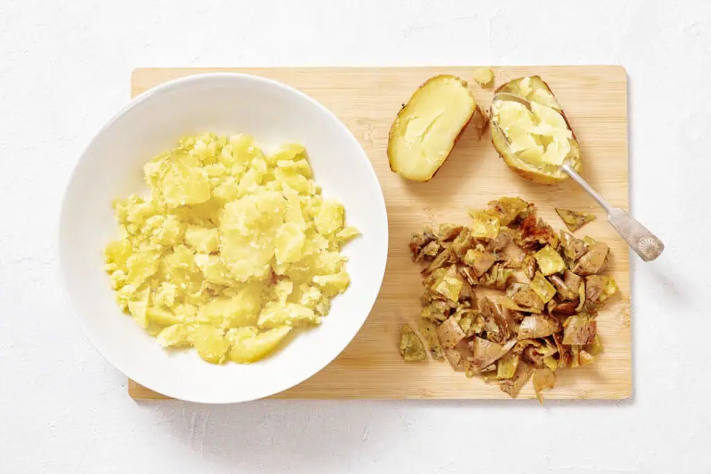 A white bowl with mashed potatoes next to diced potato skins and a halved baked potato on a wooden cutting board.