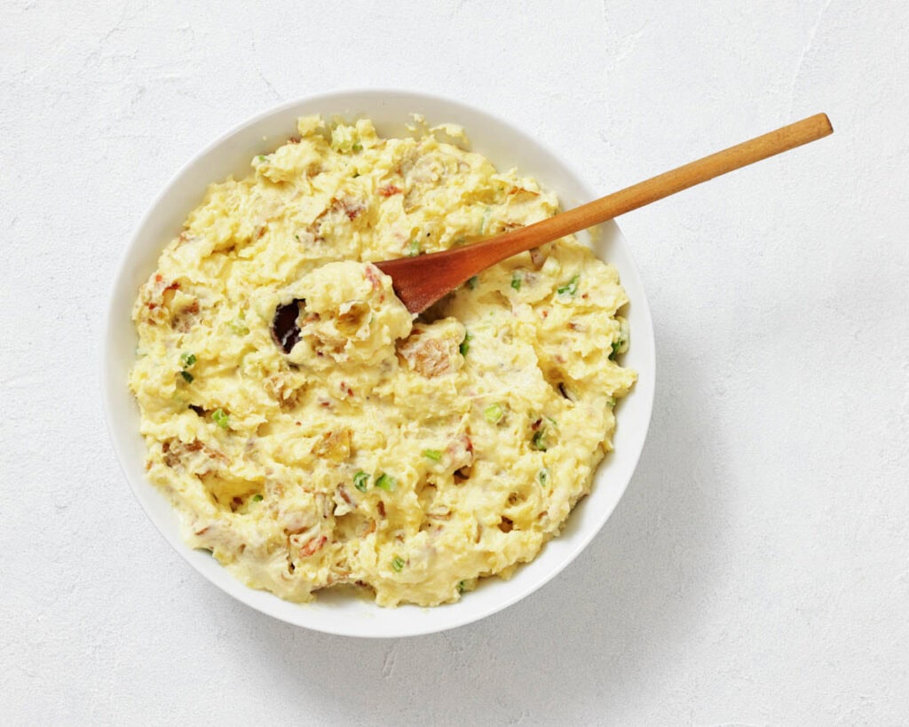 A bowl of potato salad with chopped herbs and a wooden spoon on a white surface.