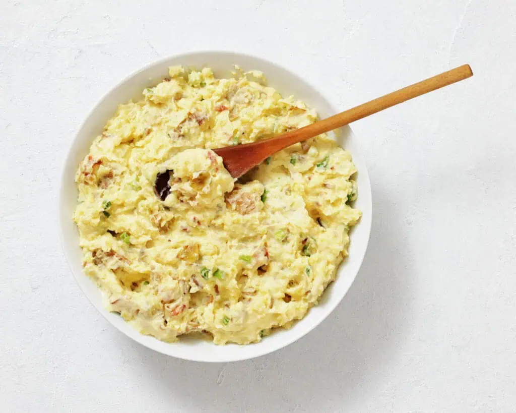 A bowl of potato salad with chopped herbs and a wooden spoon on a white surface.