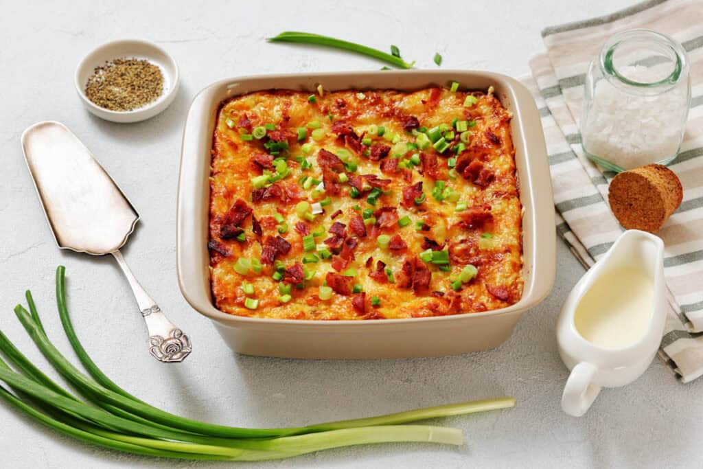 A baked casserole topped with chopped green onions and bacon bits, surrounded by fresh green onions, a dish of pepper, sea salt, bread, and a small jug of cream.