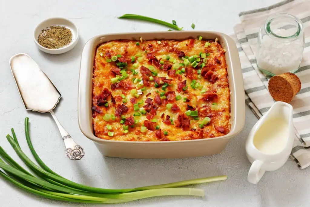 A baked casserole topped with chopped green onions and bacon bits, surrounded by fresh green onions, a dish of pepper, sea salt, bread, and a small jug of cream.