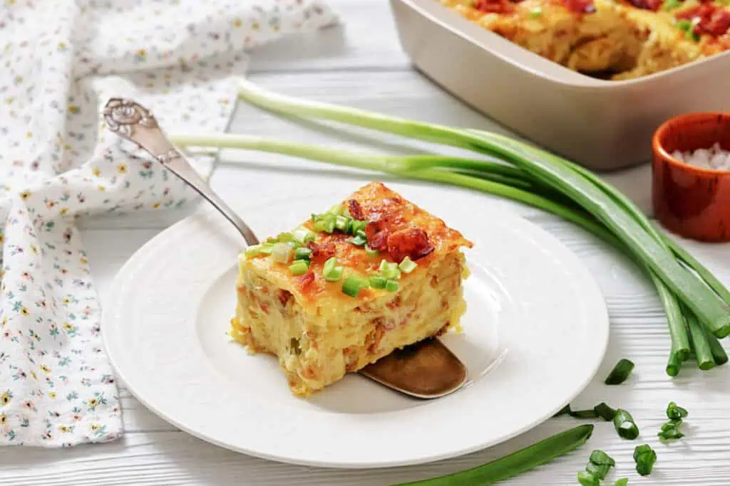 A slice of baked casserole topped with chopped green onions is served on a white plate with a spoon; green onions and a casserole dish are in the background.