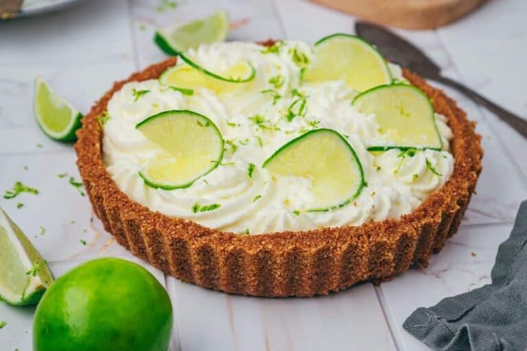 A key lime pie with a graham cracker crust, topped with whipped cream, lime slices, and lime zest, sits on a marble surface with lime wedges nearby.