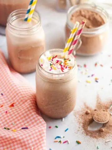 A close-up of a glass of frothy brown frozen hot chocolate, topped with white whipped cream and colorful sprinkles, with a yellow and blue striped straw, on a white countertop with a checkered napkin and another glass in the background.
