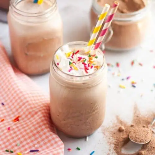 A close-up of a glass of frothy brown frozen hot chocolate, topped with white whipped cream and colorful sprinkles, with a yellow and blue striped straw, on a white countertop with a checkered napkin and another glass in the background.