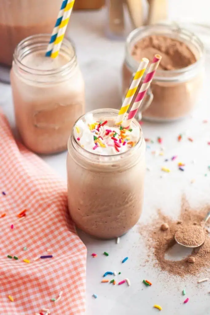 A close-up of a glass of frothy brown frozen hot chocolate, topped with white whipped cream and colorful sprinkles, with a yellow and blue striped straw, on a white countertop with a checkered napkin and another glass in the background.