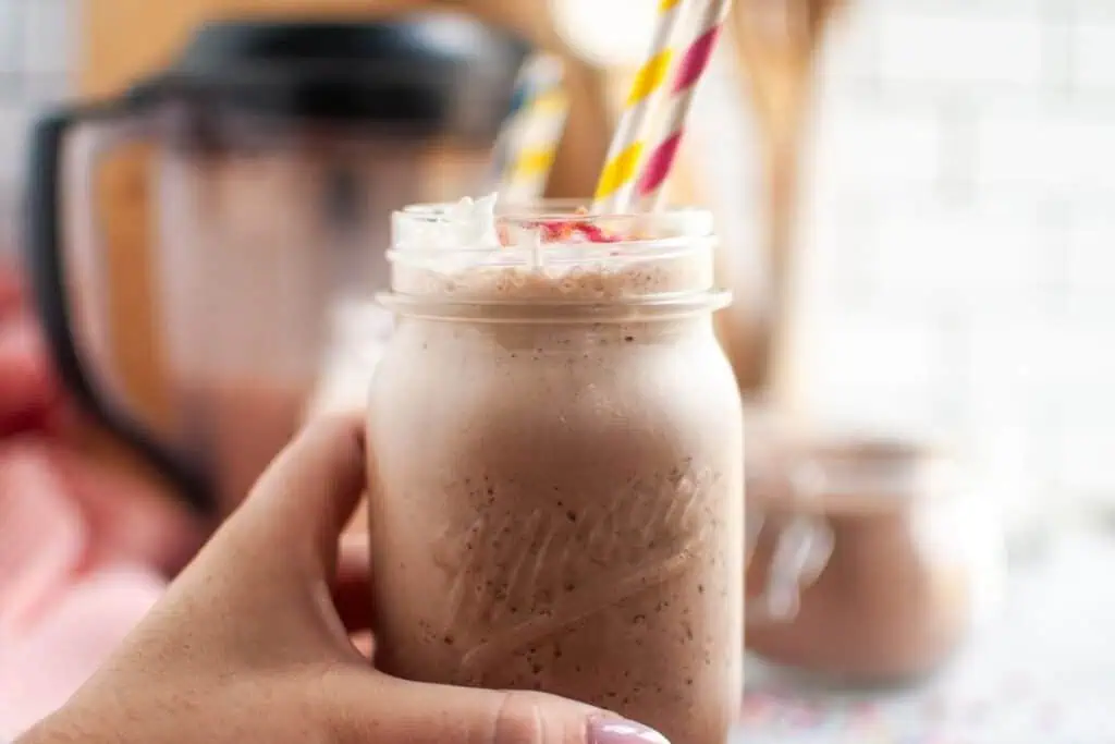 A hand with light pink fingernails holds a clear glass of frothy brown frozen hot chocolate topped with white whipped cream and colorful sprinkles.