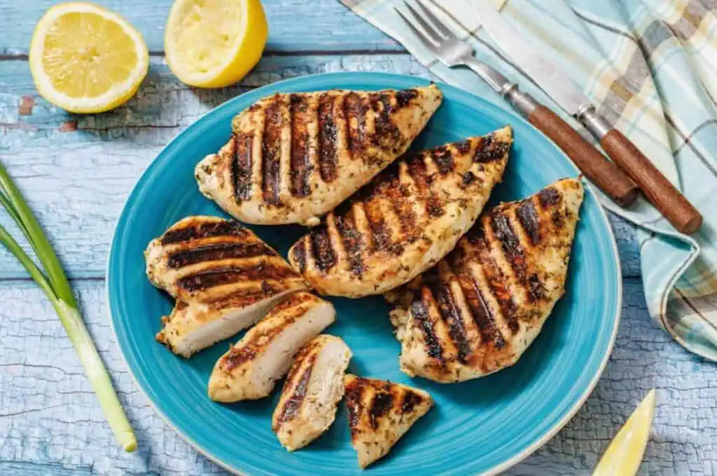Grilled chicken breasts with char marks on a blue plate, some sliced, with lemon halves and forks on a rustic blue wooden table.
