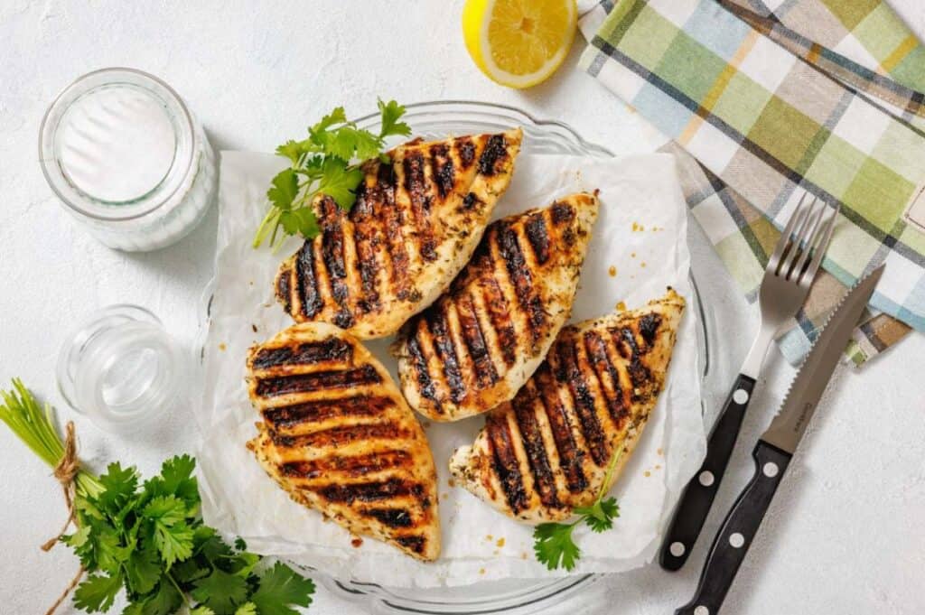 Four grilled chicken breasts on parchment paper, garnished with fresh parsley, with a lemon half, cutlery, and a checkered napkin nearby.