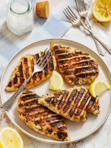 A plate with four pieces of grilled chicken breast, grill marks visible, garnished with lemon wedges; two forks, a jar of salt, and halved lemons are nearby on a table.