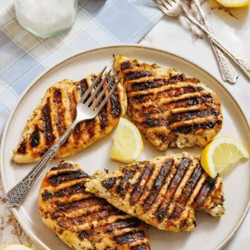 A plate with four pieces of grilled chicken breast, grill marks visible, garnished with lemon wedges; two forks, a jar of salt, and halved lemons are nearby on a table.